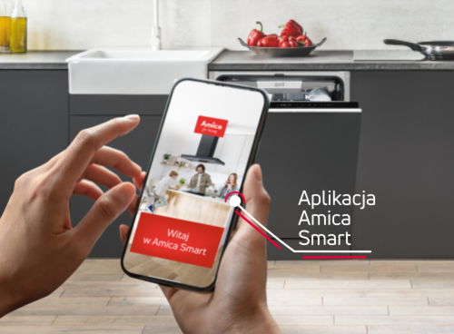Osoba trzyma smartfon z aplikacją \'Amica Smart\' i napisem \'Witaj w Amica Smart\'. W tle widać otwartą zmywarkę w zabudowie kuchennej oraz blat z czerwonymi paprykami.