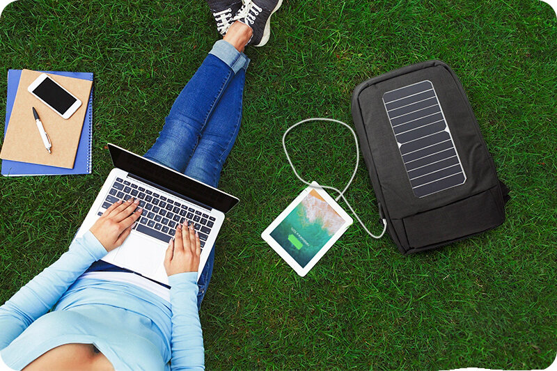 Osoba siedzi na trawie z laptopem na kolanach, obok leży plecak z wbudowanym panelem solarnym i podłączonym tabletem. Obok znajdują się notatnik, telefon i długopis.