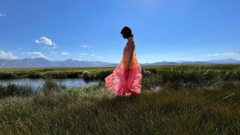Osoba w kolorowej sukni stoi na trawie obok strumienia, w tle widoczne są góry i niebo z kilkoma chmurami.