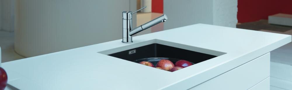 Na obrazie widoczny jest zlewozmywak z wbudowaną baterią, wypełniony wodą i jabłkami. Zlewozmywak jest umieszczony w białym blacie kuchennym.