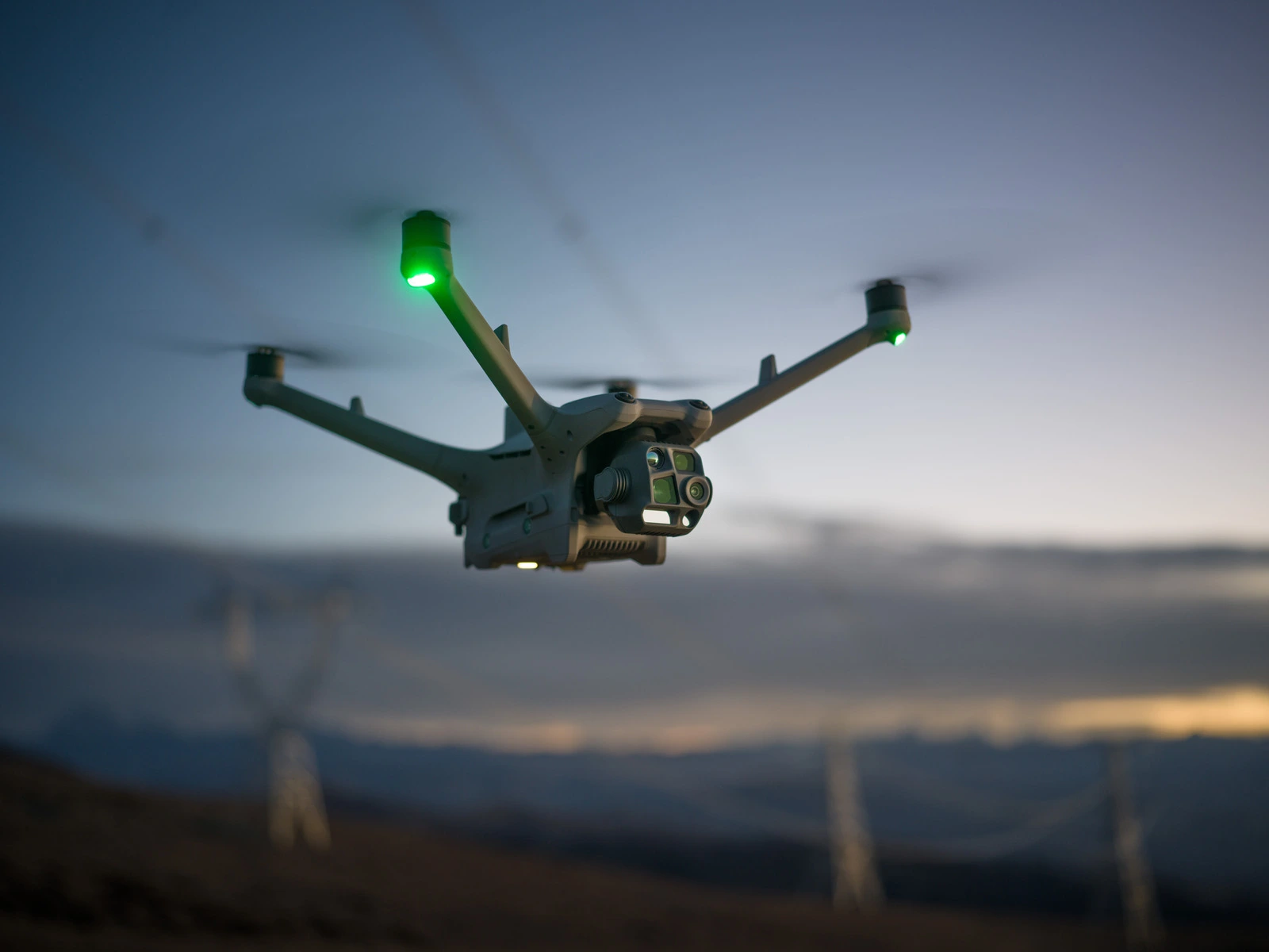 Dron z czterema wirnikami unosi się w powietrzu o zmierzchu, z widocznymi zielonymi światłami na ramionach. W tle widać rozmyte kontury krajobrazu i słupy energetyczne.