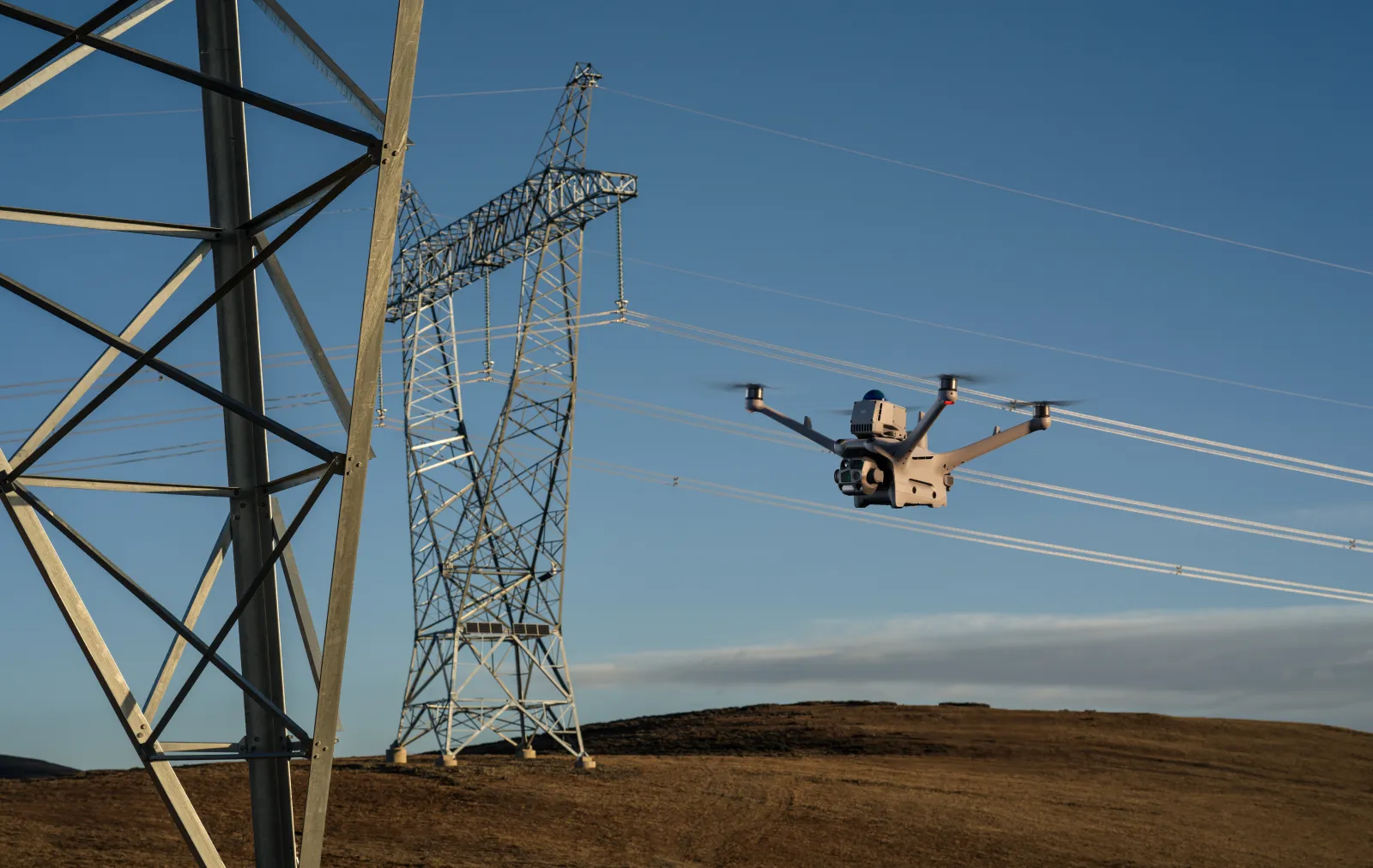 Dron latający w pobliżu konstrukcji linii wysokiego napięcia na tle nieba i pagórkowatego terenu.