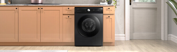 Pralka Samsung Bespoke AI™ WW11DB8B95GBU4 o pojemności 11 kg dostępna w Media Expert Czarna pralka wbudowana w ciąg szafek kuchennych o jasnobrązowym kolorze, z widocznym panelem sterowania i okrągłym bębnem. Podłoga jest drewniana, a w tle znajduje się biała ściana z drzwiami.
