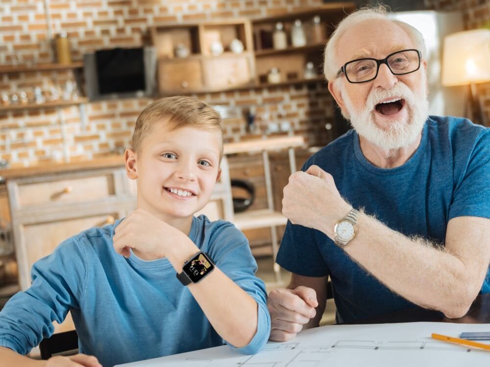 Na zdjęciu widać dwie osoby siedzące przy stole, jedna z nich nosi czarny smartwatch na nadgarstku. W tle znajduje się kuchnia z ceglaną ścianą i drewnianymi półkami.