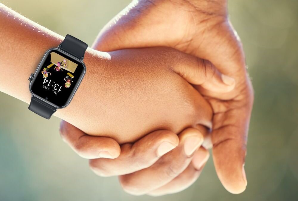 Na zdjęciu widać smartwatch z czarnym paskiem na nadgarstku dziecka, które trzyma za rękę dorosłą osobę. Na ekranie zegarka widoczna jest godzina 13:14 oraz kolorowe elementy graficzne.