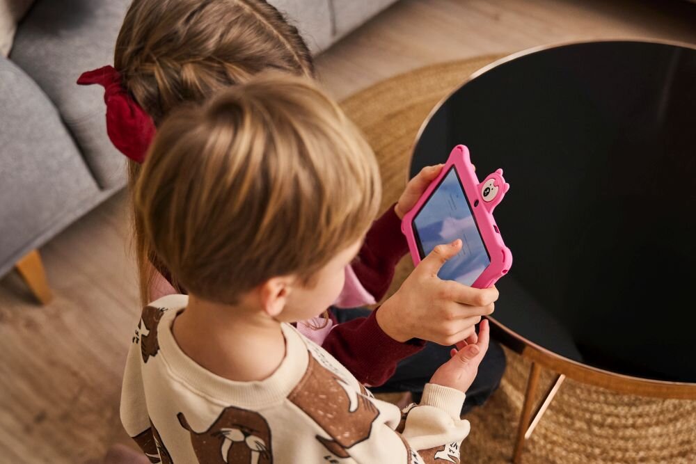 Dwoje dzieci trzyma różowy tablet z wyświetlonym obrazem. Tablet ma ochronną obudowę z uszami przypominającymi misia.
