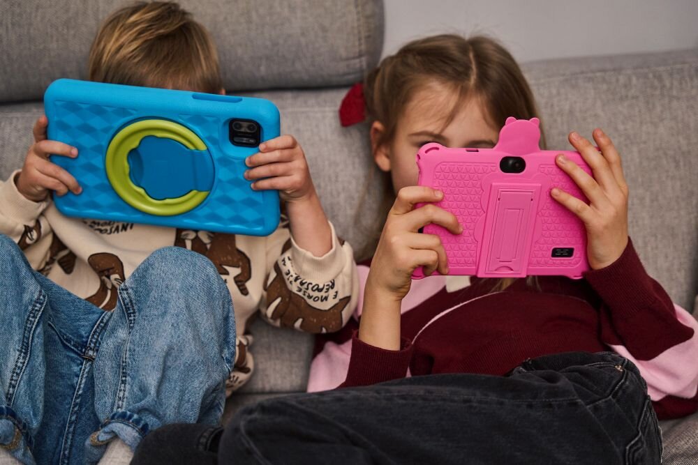 Dwoje dzieci siedzi na kanapie, każde trzyma tablet w kolorowej obudowie; jedno w niebieskiej, drugie w różowej. Obudowy mają wyraźne uchwyty i wzory, a dzieci są skupione na ekranach.