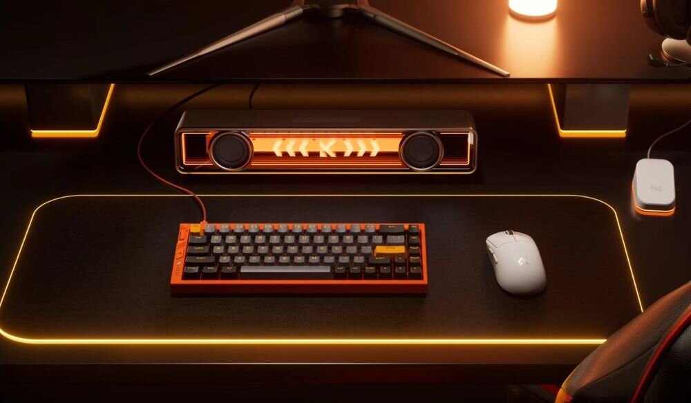 Na biurku znajduje się klawiatura z podświetleniem RGB oraz myszka, obok których leży prostokątne urządzenie z głośnikami i pomarańczowym podświetleniem. Całość jest utrzymana w ciemnej kolorystyce z pomarańczowymi akcentami świetlnymi.