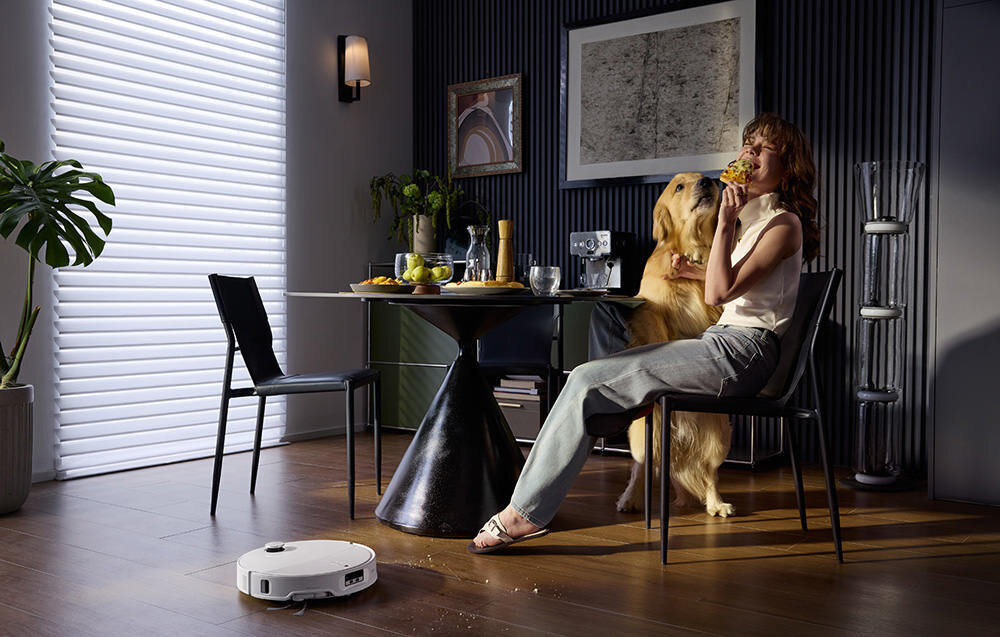 W nowoczesnym wnętrzu, na drewnianej podłodze, znajduje się biały robot sprzątający. W tle widać stół z krzesłami, na którym stoją naczynia i dekoracje.