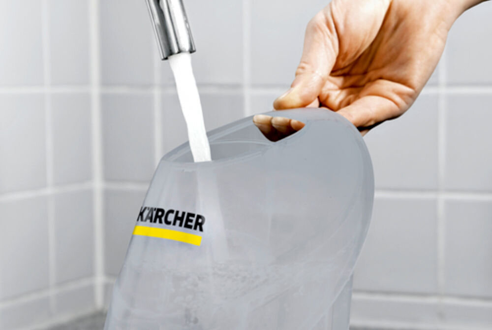 Osoba napełnia wodą przezroczysty pojemnik z logo KARCHER pod kranem. W tle widoczne są białe płytki ścienne.