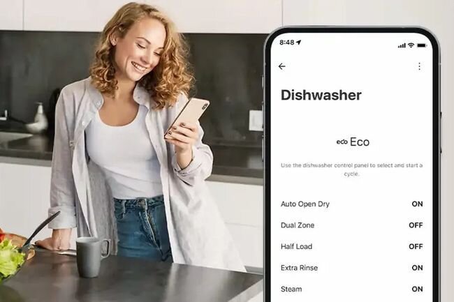 Osoba stoi w kuchni, trzymając telefon, na którym widoczny jest ekran z aplikacją do zmywarki. Na ekranie widnieje tekst: \'Dishwasher\', \'Eco\', \'Auto Open Dry ON\', \'Dual Zone OFF\', \'Half Load OFF\', \'Extra Rinse ON\', \'Steam ON\'.