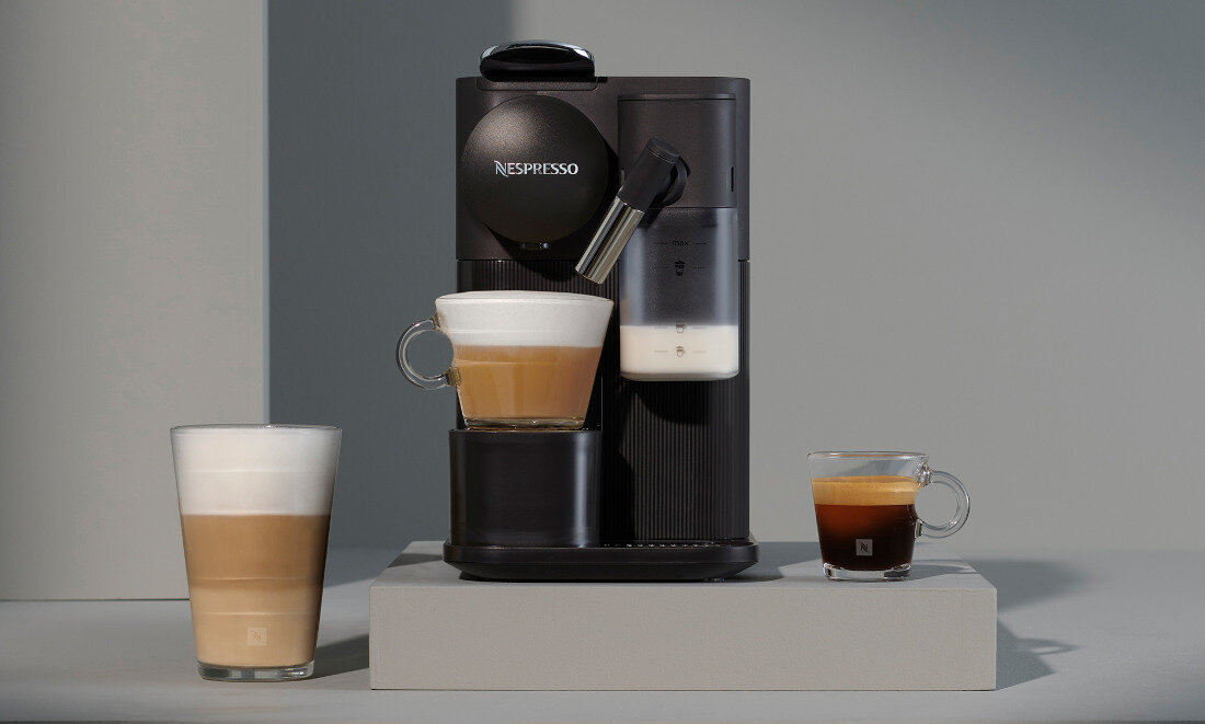 Na zdjęciu znajduje się czarny ekspres do kawy z logo Nespresso, obok którego stoją trzy filiżanki z różnymi rodzajami kawy. Ekspres ma widoczny pojemnik na mleko i dyszę do spieniania.
