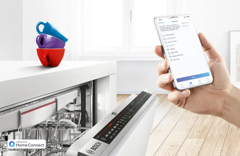 Zmywarka z otwartymi drzwiami, na których widoczne jest logo Bosch, obok osoba trzymająca smartfon z aplikacją. Na blacie stoją trzy kolorowe kubki, a w rogu widnieje logo \'Home Connect\'.