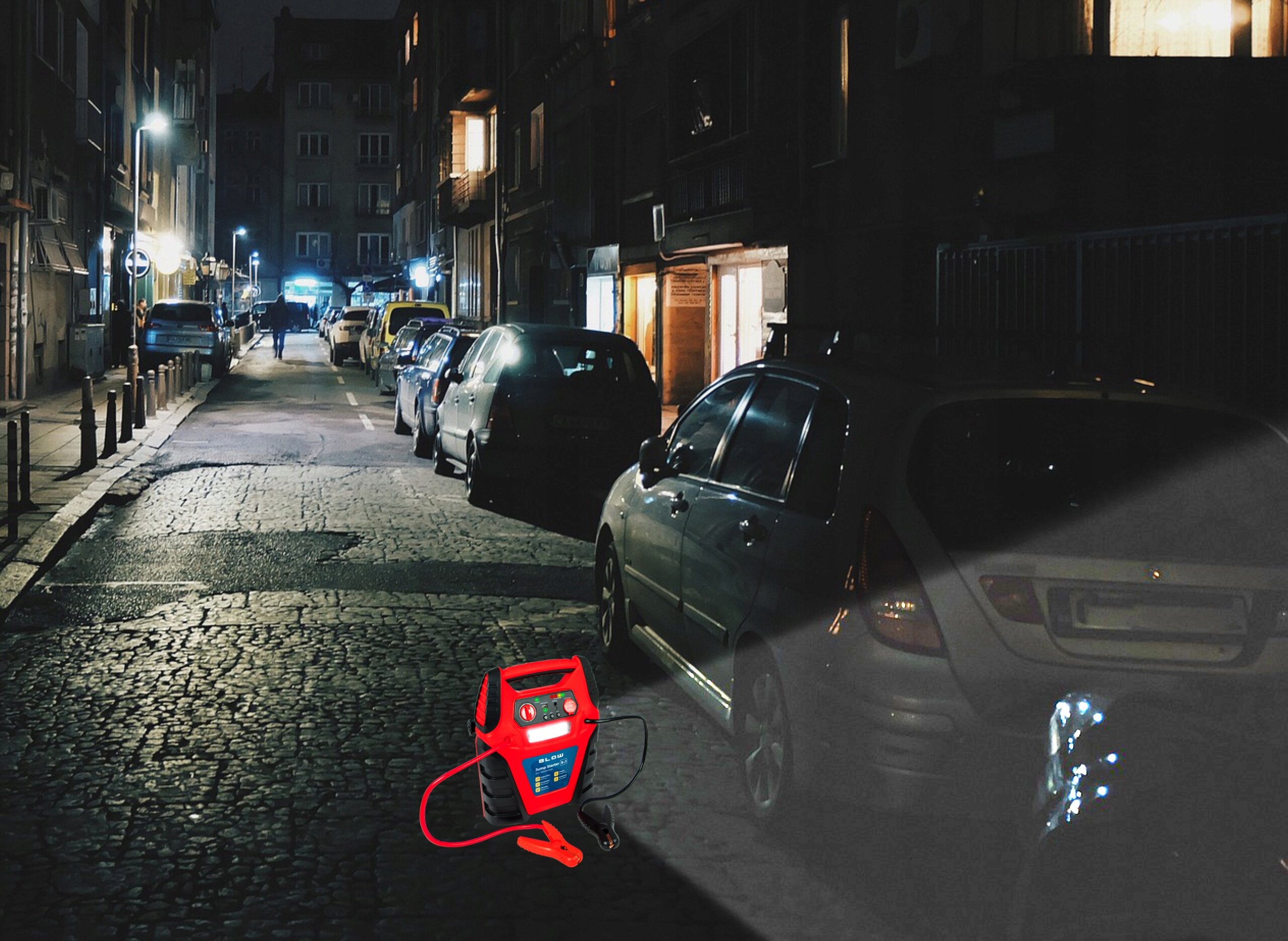 Na nocnej ulicy stoi zaparkowany samochód, a obok niego znajduje się czerwony urządzenie wielofunkcyjne z kablami i wyświetlaczem. Urządzenie emituje światło, które oświetla część pojazdu.