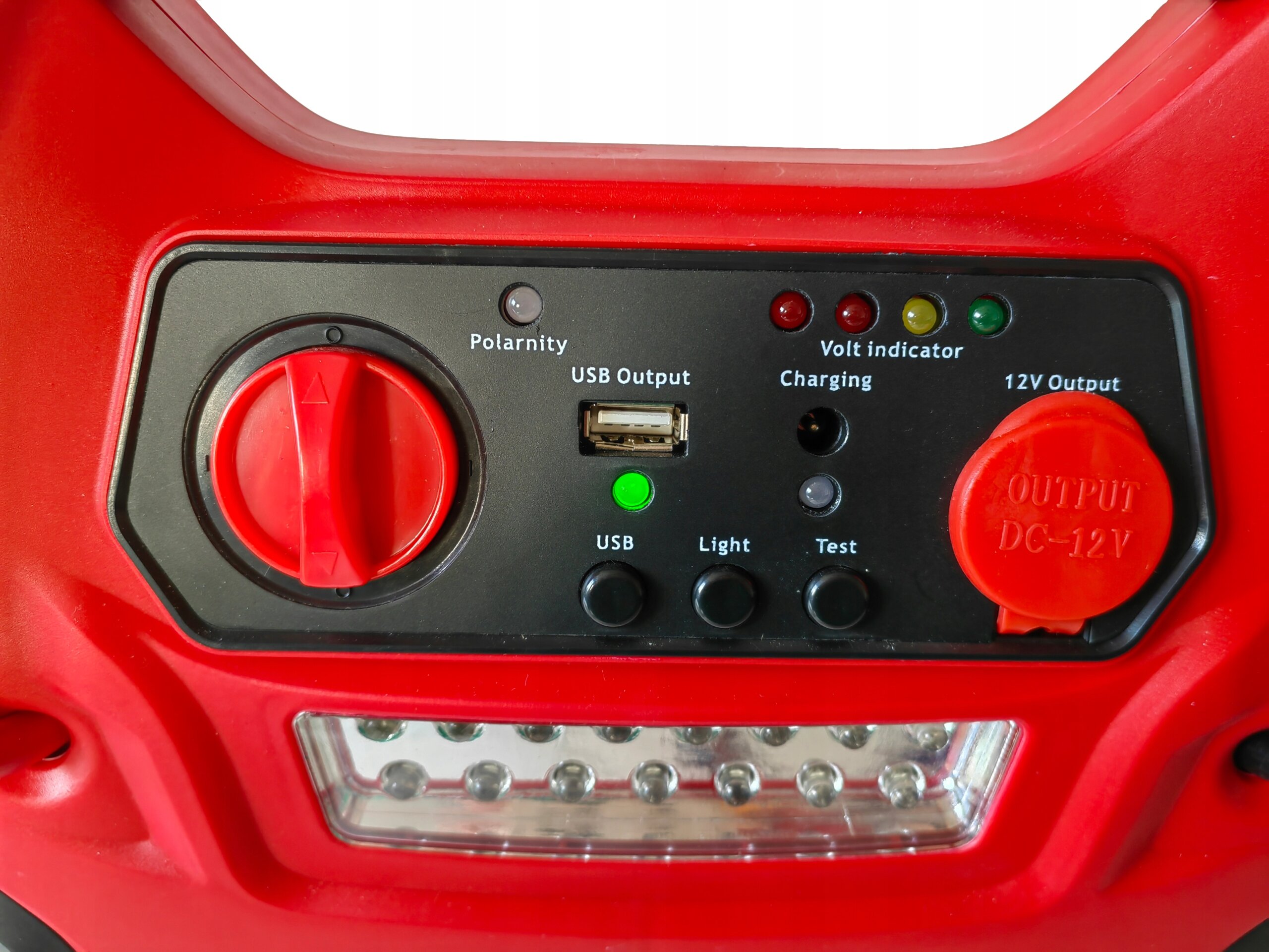Czerwony panel urządzenia z różnymi wskaźnikami i przyciskami, w tym USB Output, Volt indicator, Charging oraz 12V Output. Widoczne są także przyciski USB, Light i Test oraz napis \'OUTPUT DC-12V\'.