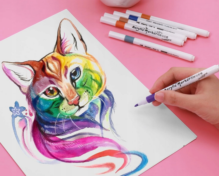Na różowym tle znajduje się kartka z kolorowym rysunkiem kota, obok której leży kilka markerów z napisem \'Acrylic Marker\'. Ręka trzyma jeden z markerów, dodając fioletowy kolor do rysunku.
