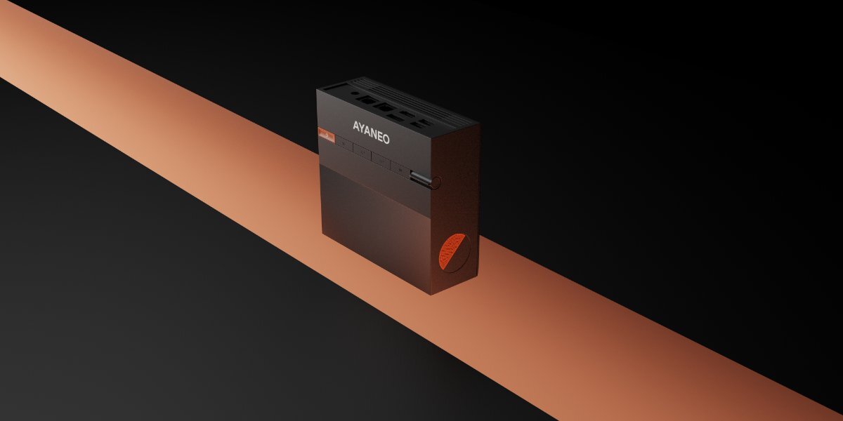 Czarny mini PC AYANEO z widocznym logo \'AYANEO\' na górnej części obudowy, umieszczony na pomarańczowym tle. Obok znajduje się pomarańczowy element dekoracyjny w kształcie liścia.