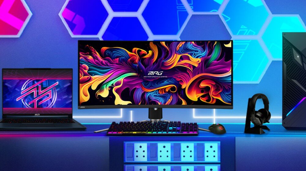 Na biurku znajduje się monitor z kolorowym, abstrakcyjnym wzorem na ekranie, obok laptopa i komputera stacjonarnego. Na monitorze widoczne jest logo \'MPG\' oraz napis \'Gaming Series\'.