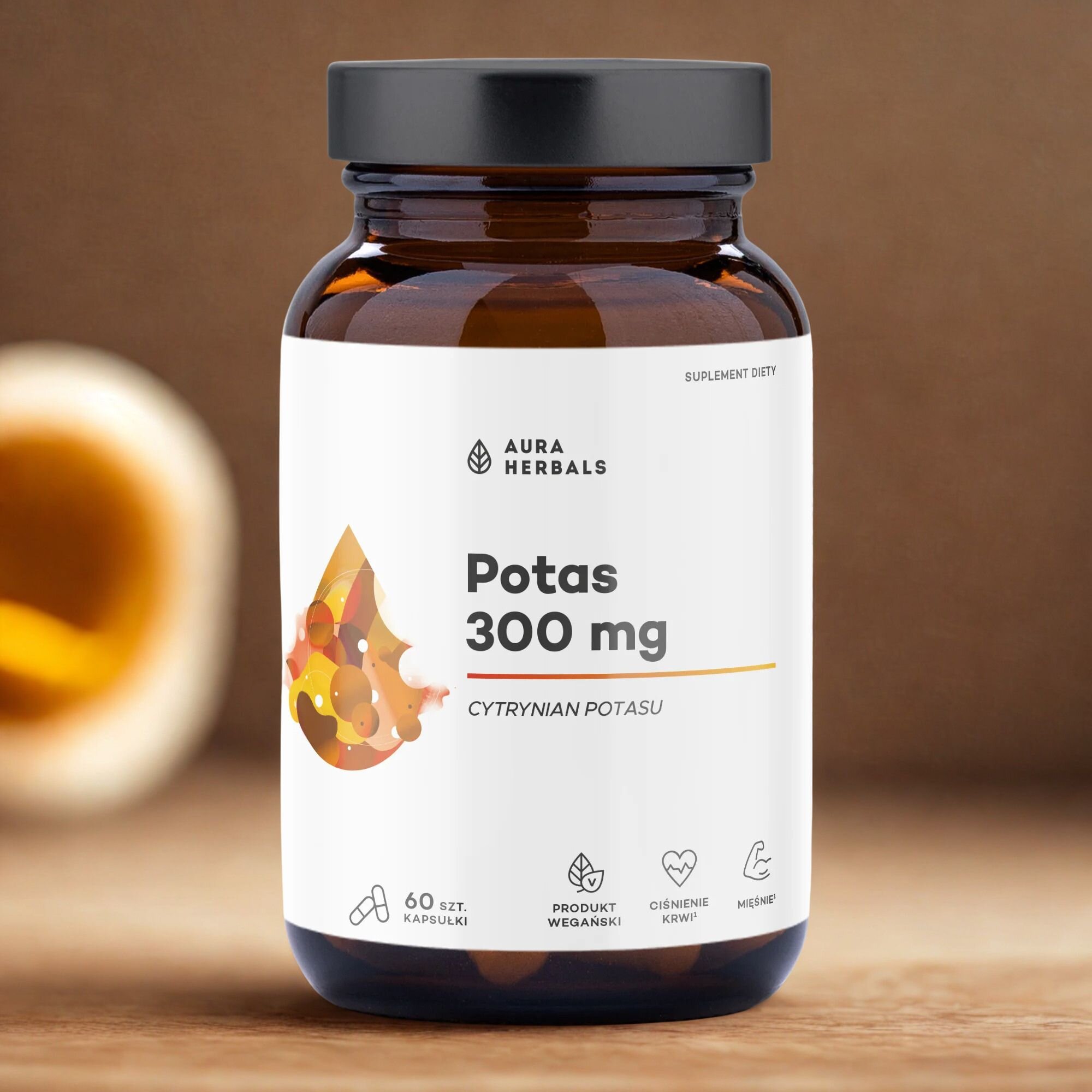 Na obrazie widoczna jest brązowa butelka z etykietą \'Aura Herbals Potas 300 mg Cytrynian Potasu\'. Na dole etykiety znajdują się ikony z napisami: \'60 szt. kapsułki\', \'Produkt wegański\', \'Ciśnienie krwi\', \'Mięśnie\'.