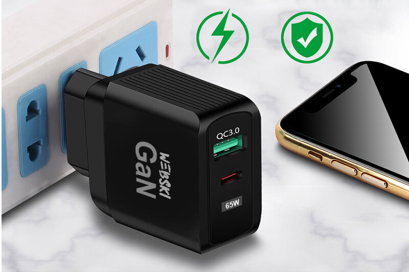 Czarna ładowarka sieciowa z dwoma portami USB, oznaczona jako \'QC3.0\' i \'65W\', podłączona do gniazdka. Obok znajduje się smartfon oraz ikony symbolizujące szybkie ładowanie i bezpieczeństwo.