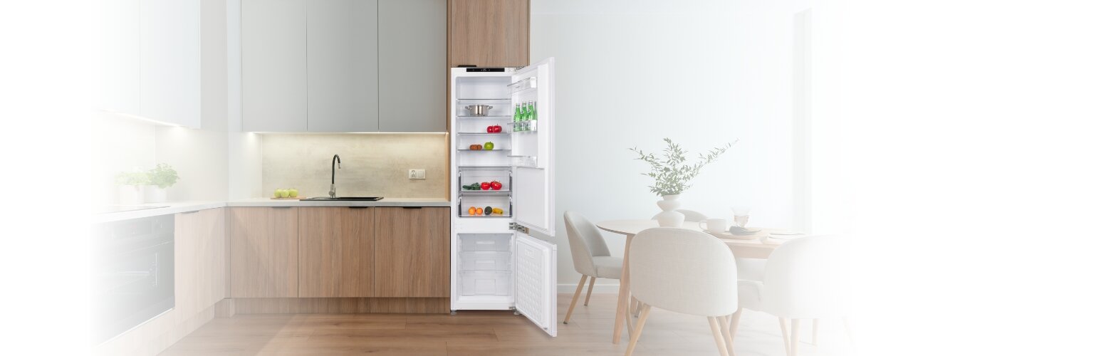 Kuchnia z otwartą lodówką w zabudowie, w której znajdują się różne produkty spożywcze. Obok lodówki stoi stół z krzesłami, a na blacie kuchennym widoczny jest zlew i płyta grzewcza.