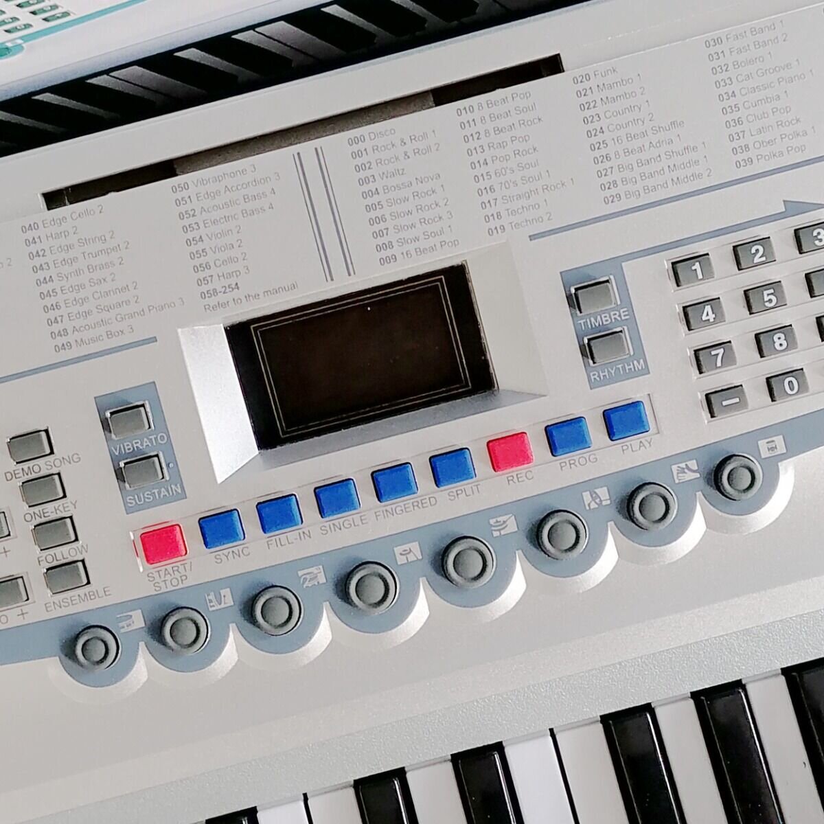 Panel sterowania elektronicznego keyboardu z wyświetlaczem i przyciskami w różnych kolorach, w tym czerwonymi i niebieskimi. Widoczne są także numery i nazwy różnych stylów muzycznych oraz przyciski funkcyjne takie jak \'DEMO SONG\', \'VIBRATO\', \'SUSTAIN\'.