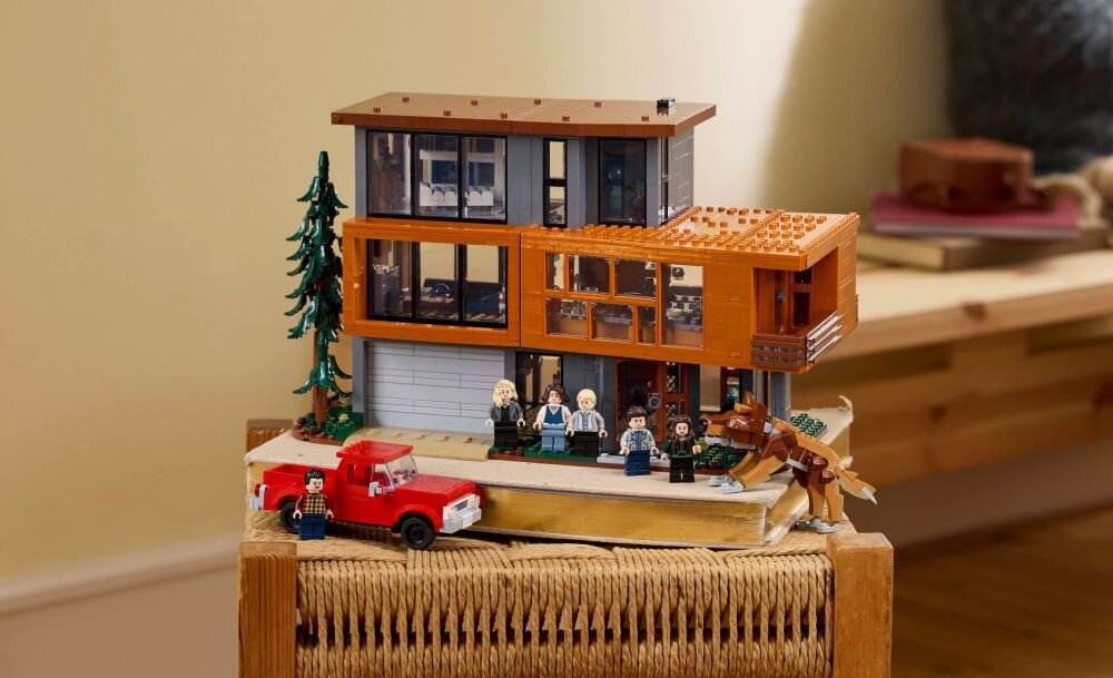 Model domu z klocków LEGO przedstawiający nowoczesny budynek z dużymi oknami i tarasem, obok którego stoi czerwona ciężarówka oraz kilka figurek. W tle widoczne są elementy wystroju wnętrza.