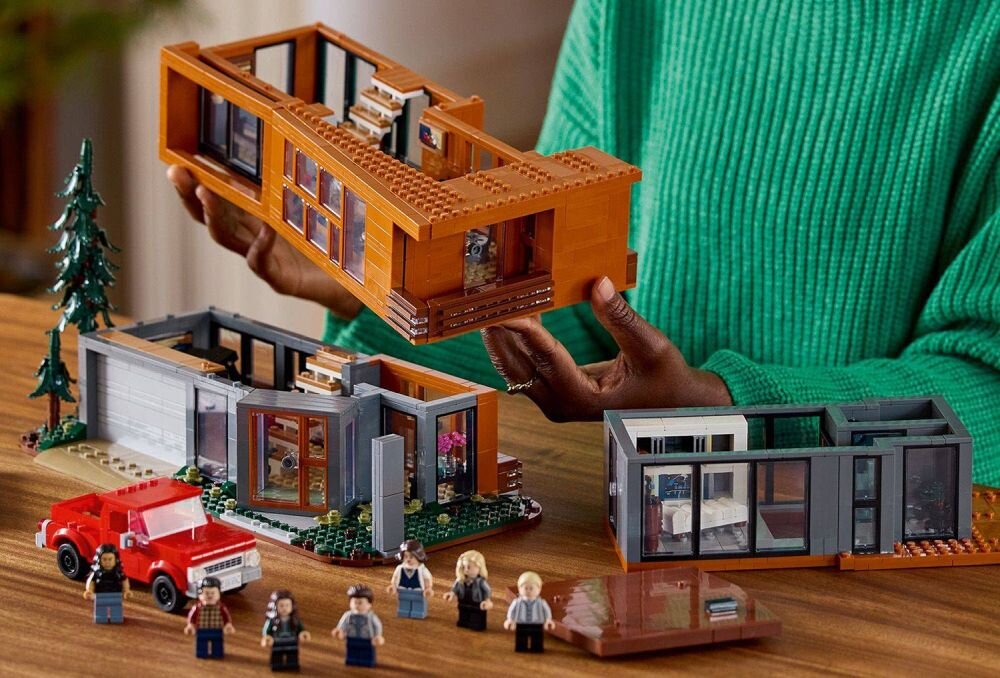 Zestaw LEGO przedstawiający dom z brązowymi i szarymi elementami oraz czerwony samochód. Przed domem znajdują się figurki postaci LEGO.