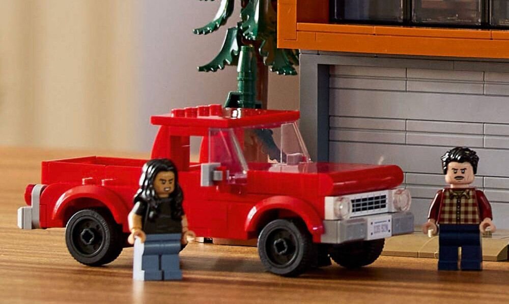 Na obrazie znajduje się czerwony samochód z klocków LEGO oraz dwie figurki LEGO stojące obok niego. W tle widoczna jest część budynku z szarymi cegłami i pomarańczowym elementem.