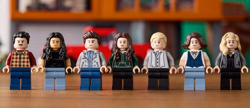 Na drewnianym stole stoi siedem figurek LEGO przedstawiających postacie z różnymi fryzurami i ubraniami. W tle widoczne są rozmyte elementy wnętrza.