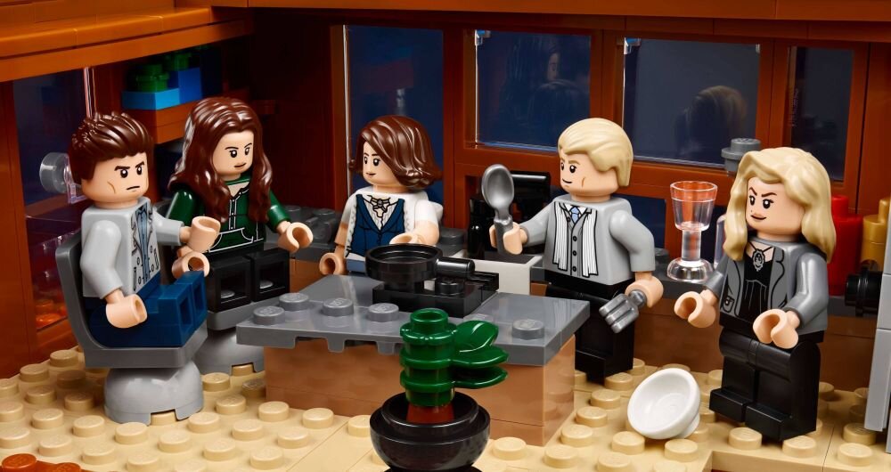 Zestaw LEGO przedstawiający pięć figurek w salonie z meblami i rośliną na środku. Figurka trzyma kieliszek, a inna łyżkę, na stole znajdują się naczynia.