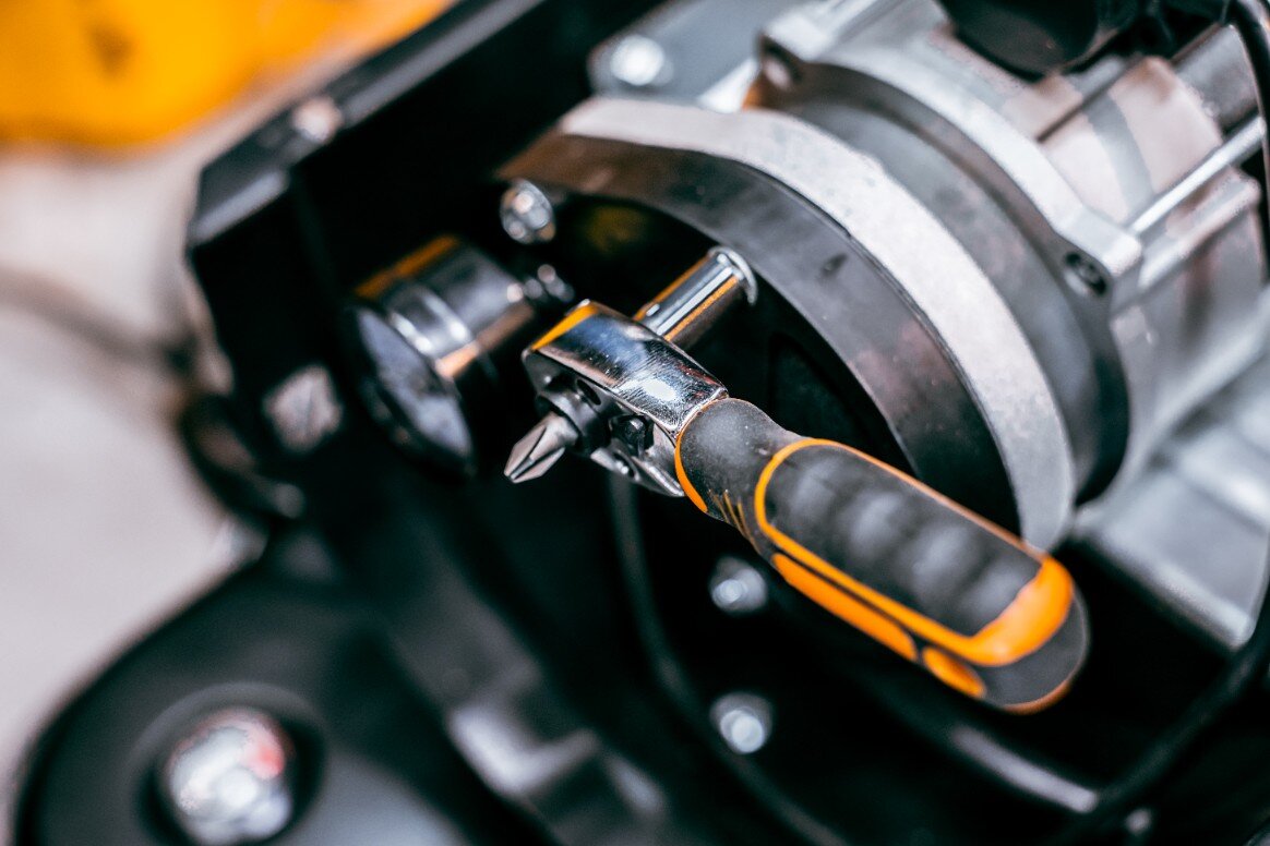 Na obrazie widoczna jest grzechotka z pomarańczowo-czarnym uchwytem, zamocowana na metalowej konstrukcji. Narzędzie jest częścią większego mechanizmu.