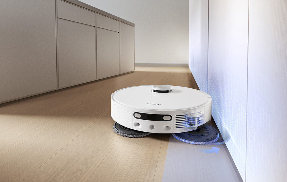 Na podłodze znajduje się biały robot sprzątający z widocznymi szczotkami i panelem sterowania. Na obudowie widoczny jest napis \'DREAME\'.