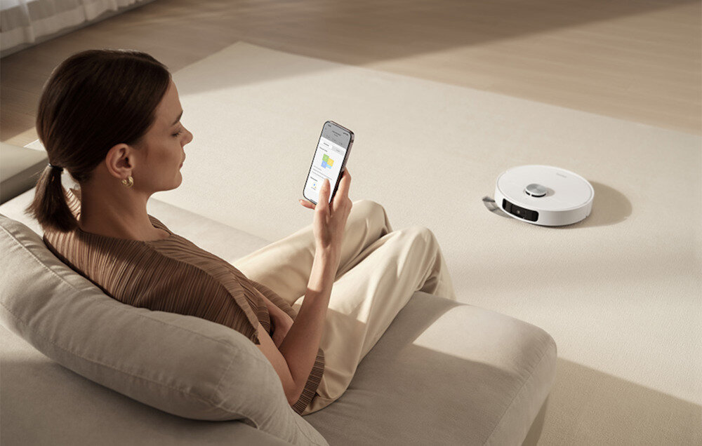 Osoba siedzi na kanapie, trzymając w ręku smartfon z widocznym ekranem. Na podłodze znajduje się biały robot sprzątający.