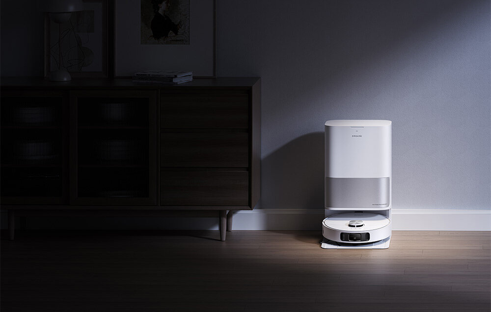 Na podłodze w ciemnym pomieszczeniu stoi biały robot sprzątający z bazą ładującą. Na urządzeniu widoczne jest logo \'Dreame\'.