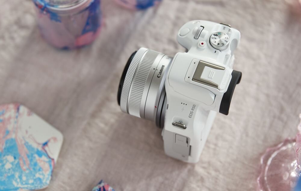 Biały aparat bezlusterkowy z widocznym logo Canon i oznaczeniem EOS R50, leży na jasnym tle. Obok aparatu znajdują się kolorowe elementy dekoracyjne.