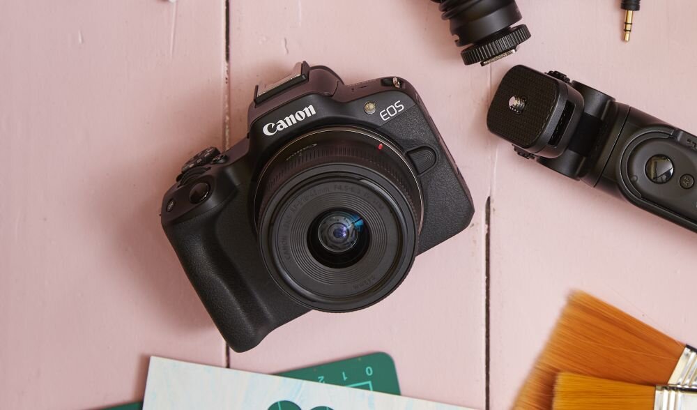 Na obrazie widoczny jest aparat fotograficzny z logo \'Canon\' i oznaczeniem \'EOS\'. Obok aparatu znajdują się akcesoria fotograficzne oraz pędzle na różowym tle.