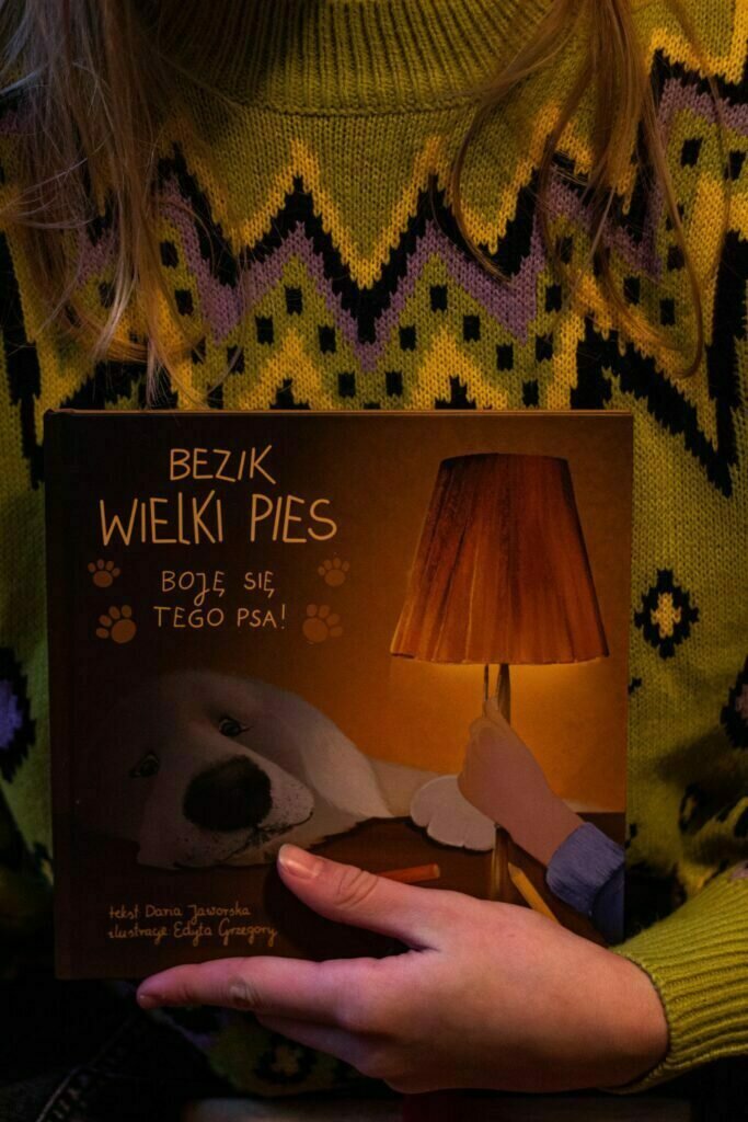 Osoba trzyma książkę z okładką przedstawiającą psa i lampę, z napisem \'Bezik Wielki Pies – Boję się tego psa!\'.