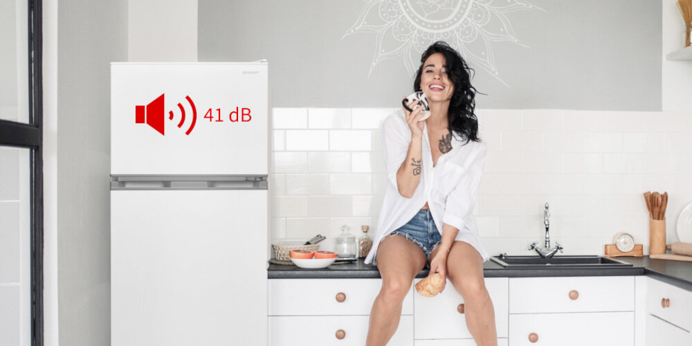Biała lodówka z symbolem głośnika i napisem \'41 dB\' na drzwiach. Kobieta siedzi na blacie kuchennym obok lodówki, trzymając kubek.