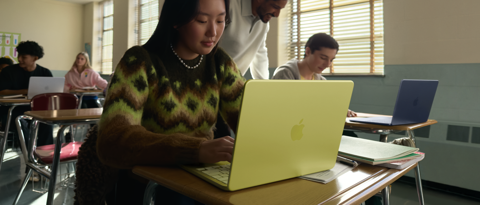 Osoba siedzi przy biurku w klasie, używając żółtego laptopa z logo w kształcie jabłka. W tle widać inne osoby przy biurkach oraz okna z żaluzjami.
