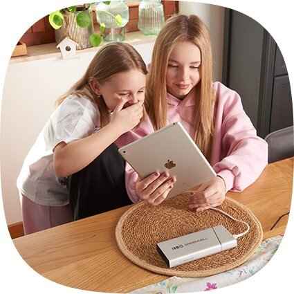 Na stole leży srebrny powerbank z napisem \'GREENCELL\', podłączony do tabletu trzymanego przez dwie osoby.