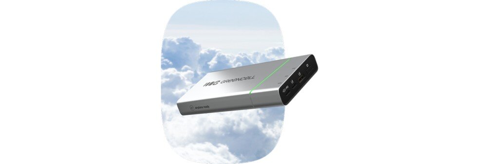 Srebrny powerbank z logo \'GREENCELL\' unosi się na tle nieba z chmurami. Na urządzeniu widoczne są porty USB i wskaźnik LED.