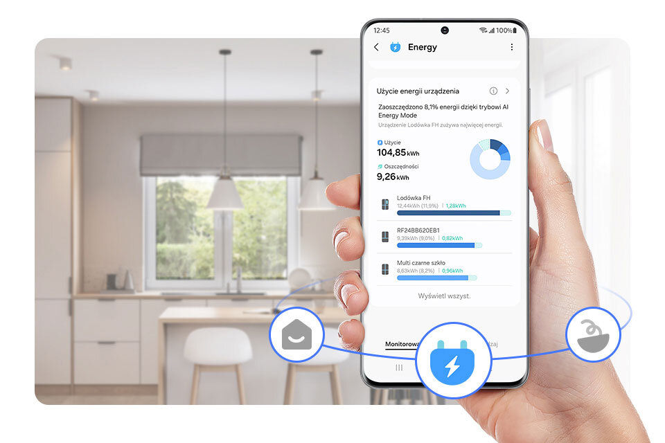 Osoba trzyma smartfon z aplikacją pokazującą zużycie energii urządzeń, w tle widoczna jest nowoczesna kuchnia. Na ekranie telefonu widnieje tekst: \'Energy\', \'Użycie energii urządzenia\', \'104,85 kWh\', \'9,26 kWh\', \'Lodówka FH\', \'RF28BB6200B1\', \'Multi czarne szkło\'.