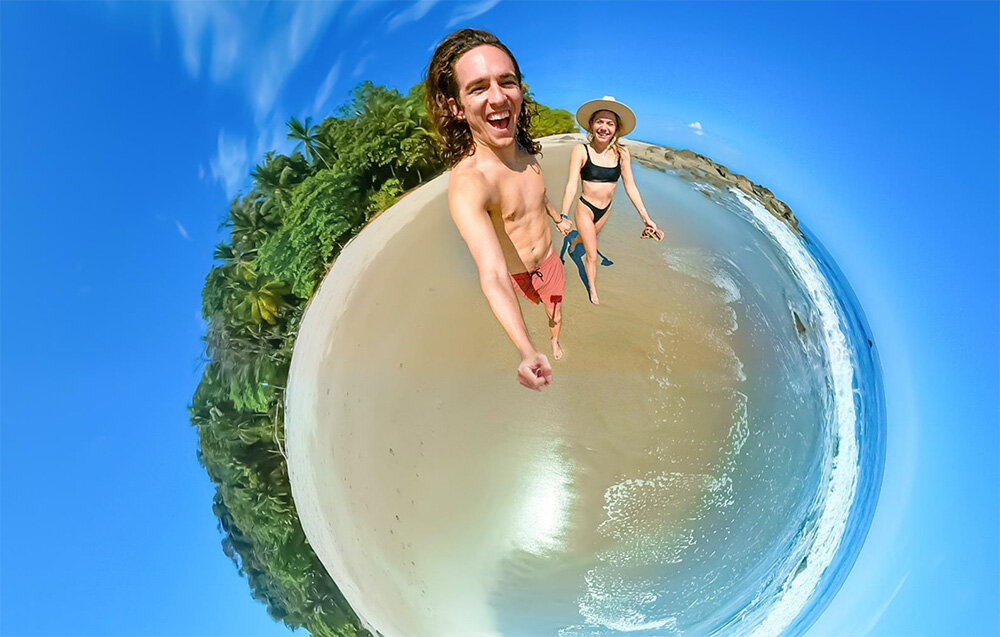 Zdjęcie przedstawia efekt sferyczny z plażą, oceanem i palmami w tle. Dwie osoby uśmiechają się, trzymając kamerę na wysięgniku.