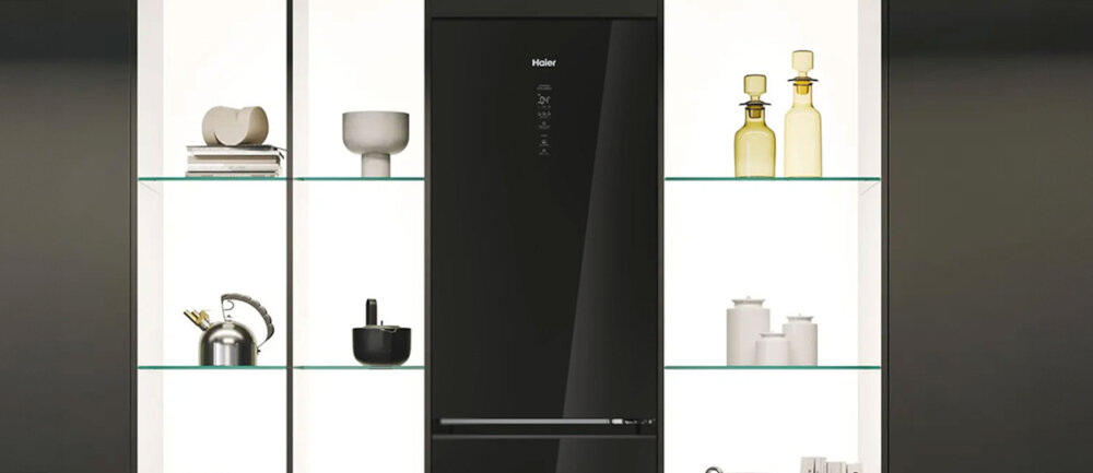 Czarna lodówka z logo \'Haier\' umieszczona w zabudowie, otoczona półkami z dekoracjami i naczyniami. Na drzwiach lodówki widoczny jest panel dotykowy z ikonami.