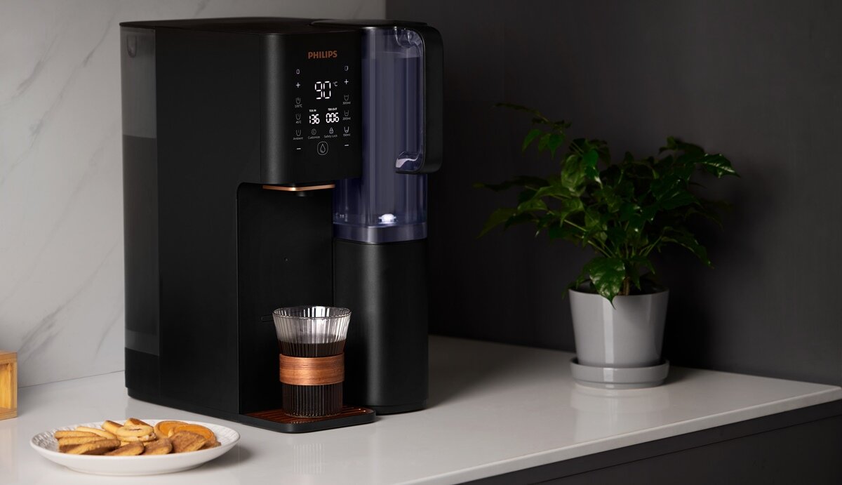 Czarny dyspenser do wody marki PHILIPS z wyświetlaczem cyfrowym, obok którego stoi szklanka na podstawce. Na blacie znajduje się również talerz z ciastkami i doniczka z rośliną.