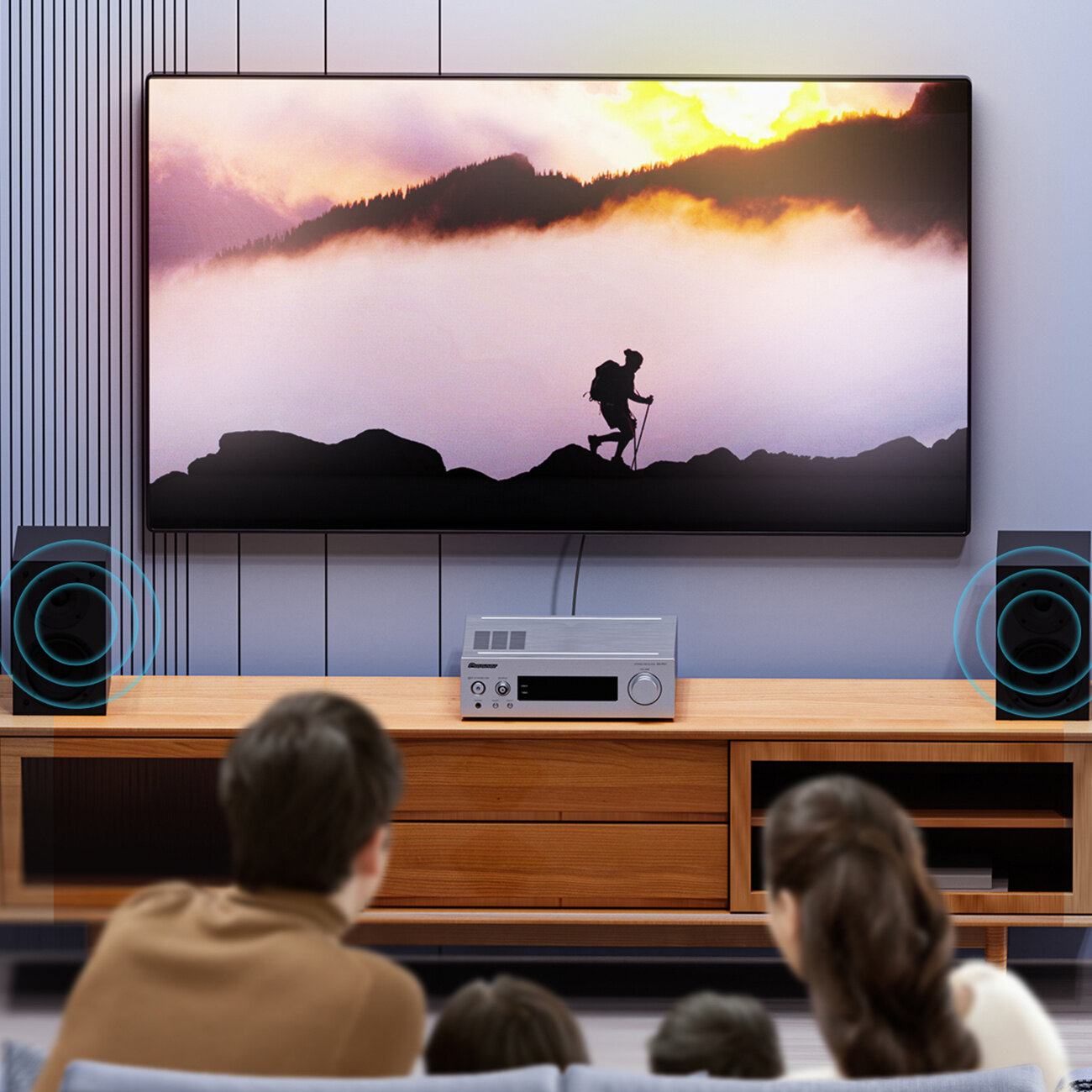 Na obrazie widać telewizor wyświetlający sylwetkę wędrowca na tle gór i zachodzącego słońca. Przed telewizorem znajduje się drewniana szafka z urządzeniem audio i głośnikami po bokach.