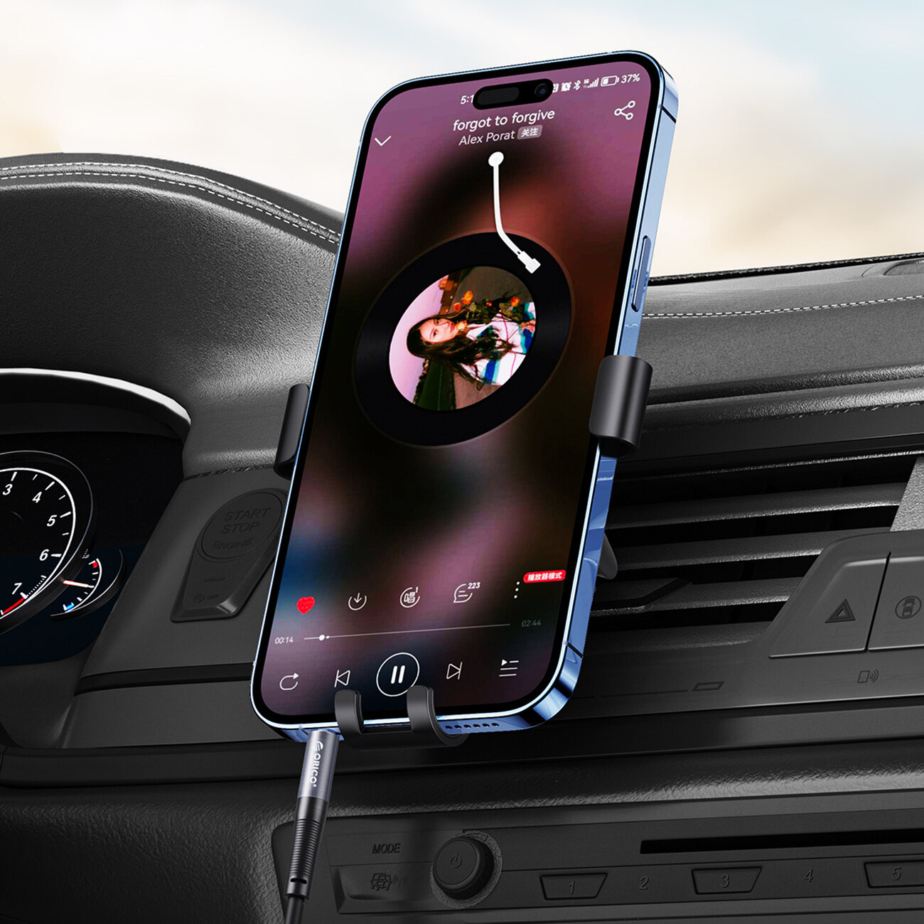 Smartfon zamocowany w uchwycie samochodowym, z ekranem wyświetlającym odtwarzacz muzyki z utworem \'forgot to forgive\' Alex Porat. Do telefonu podłączony jest kabel audio z widocznym logo \'Orico\'.