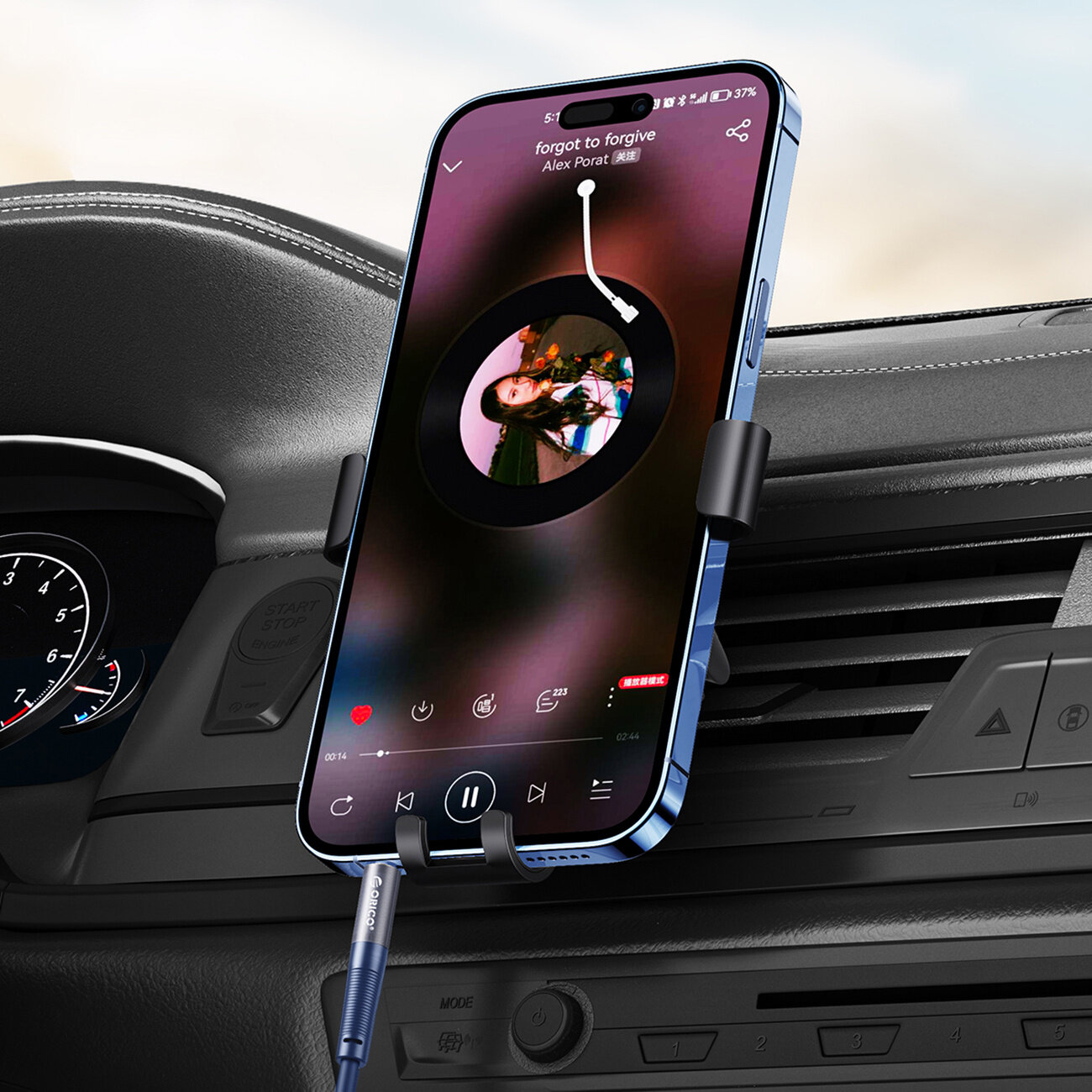 Smartfon zamocowany w uchwycie samochodowym z odtwarzaczem muzyki na ekranie, podłączony niebieskim kablem audio. Na ekranie widoczny jest tekst: \'forgot to forgive Alex Porat\'.