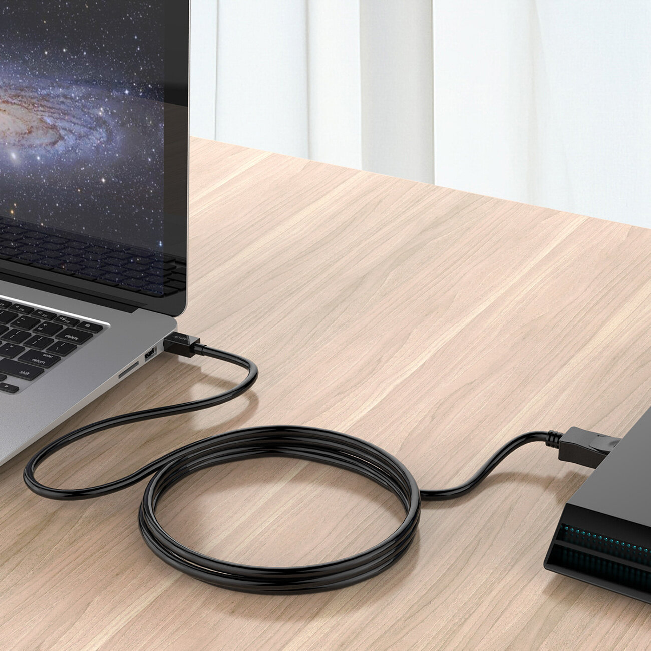 Na drewnianym stole znajduje się laptop z podłączonym czarnym kablem, który prowadzi do innego urządzenia. Kabel jest zwinięty w pętlę na blacie.